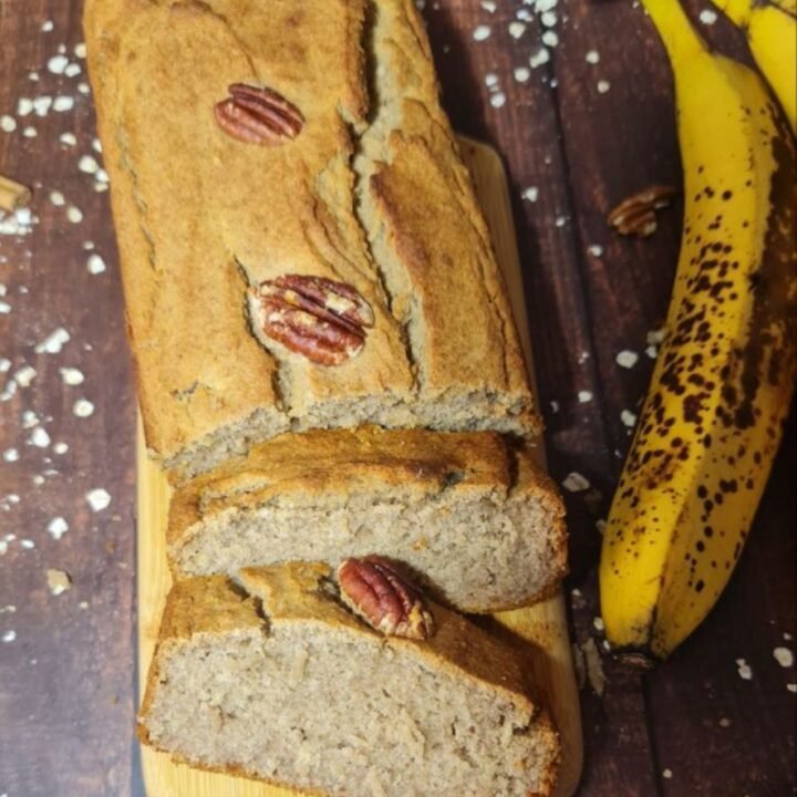 Vegan banana bread loaf topped with pecans, sliced on a wooden board with ripe bananas and scattered oats in the background.