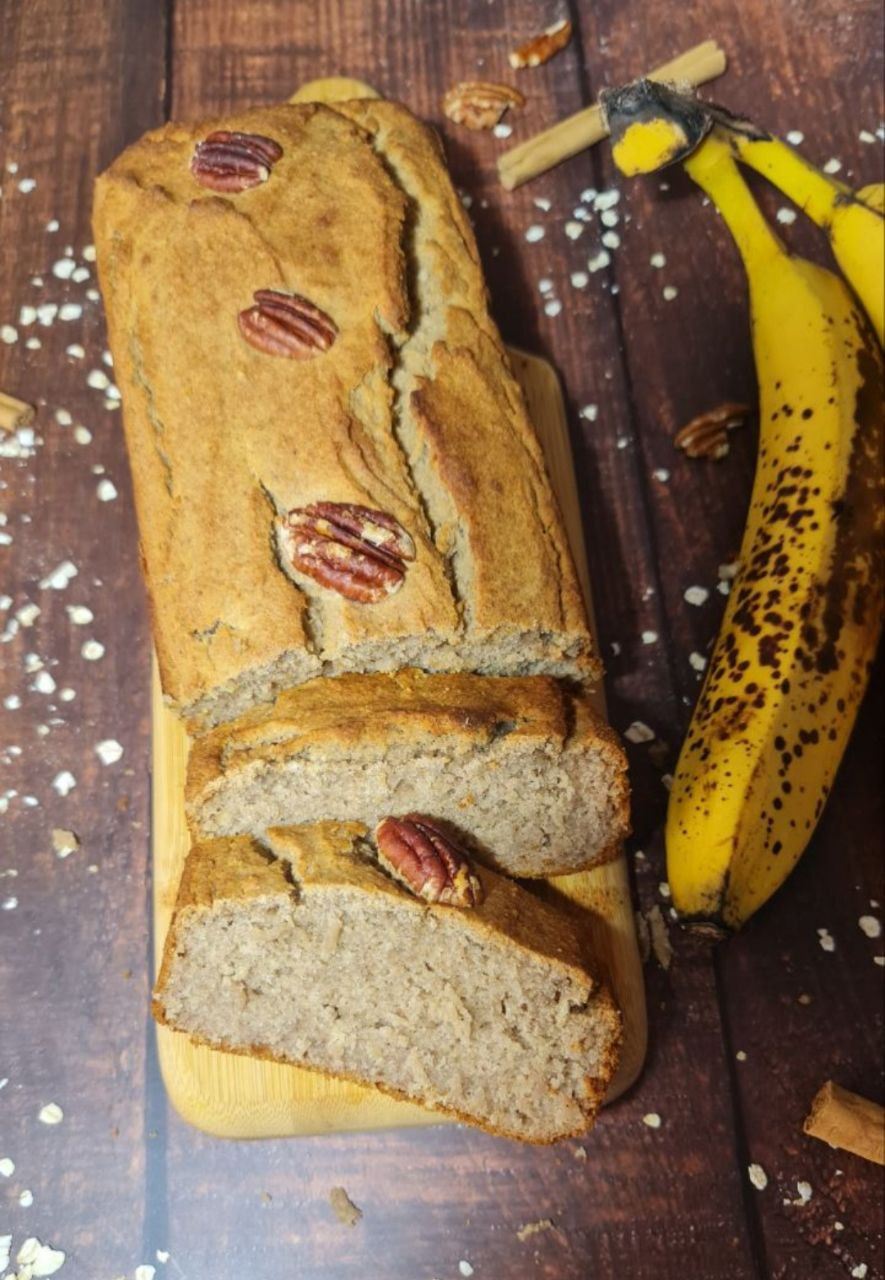 Vegan banana bread loaf topped with pecans, sliced on a wooden board with ripe bananas and scattered oats in the background.