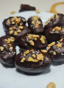 Chocolate-covered dates sprinkled with crushed peanuts and sea salt on a white plate, garnished with chocolate pieces and peanuts.
