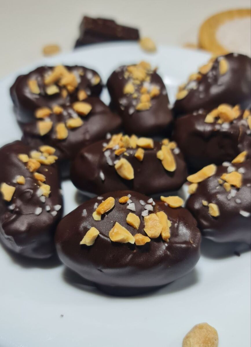 Chocolate-covered dates sprinkled with crushed peanuts and sea salt on a white plate, garnished with chocolate pieces and peanuts