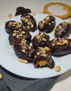 Close-up of chocolate-covered dates with peanut butter filling, sprinkled with crushed peanuts, displayed on a white plate.