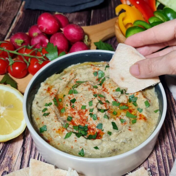 Freshly made baba ganoush in a round bowl garnished with parsley and paprika, surrounded by colorful raw vegetables like carrots, radishes, and green bell peppers, and served with pita bread on a rustic wooden table.