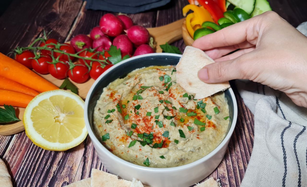 Freshly made baba ganoush in a round bowl garnished with parsley and paprika, surrounded by colorful raw vegetables like carrots, radishes, and green bell peppers, and served with pita bread on a rustic wooden table.