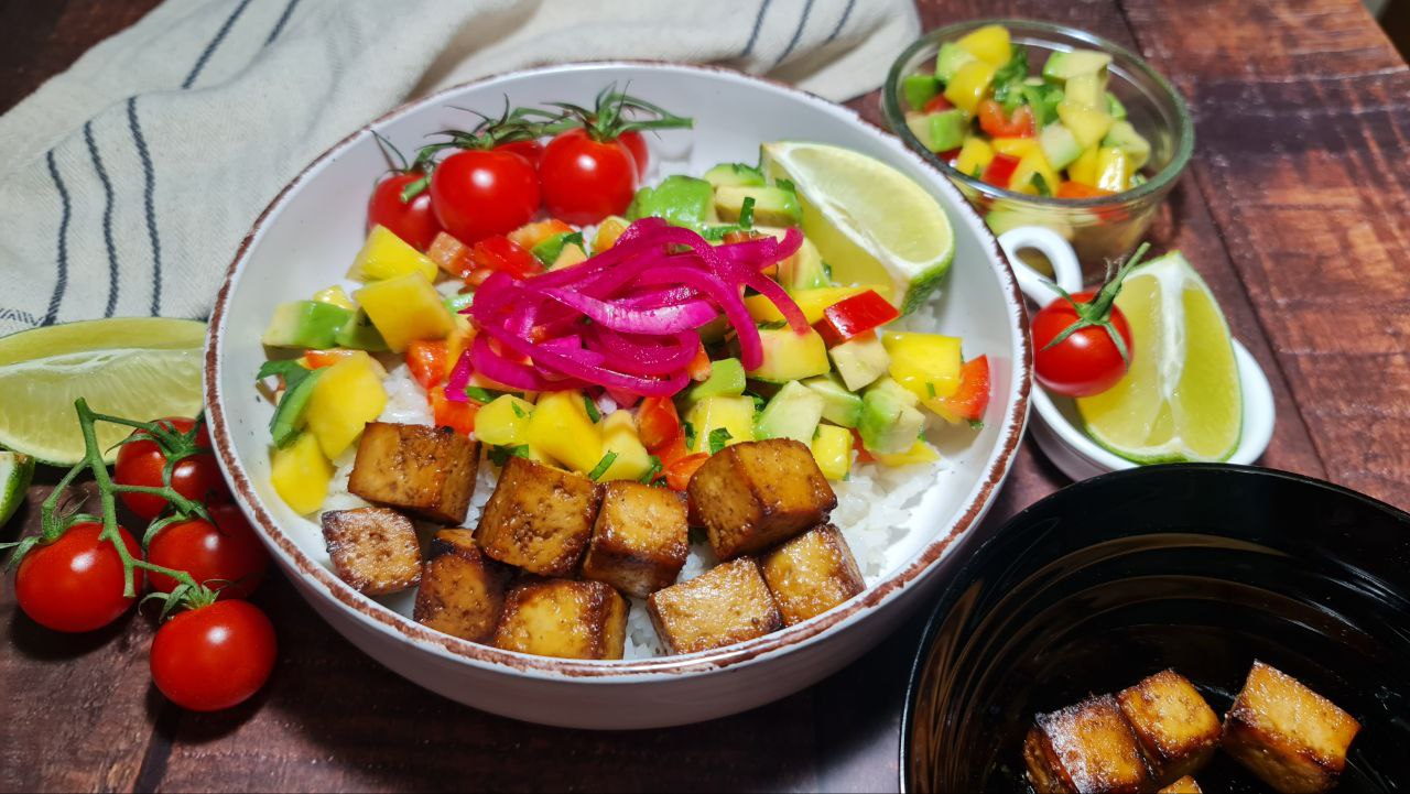 Colorful tropical tofu rice bowl with bright pink pickled onions, avocado mango salsa, crispy tofu, and fresh cherry tomatoes, garnished with lime wedges.