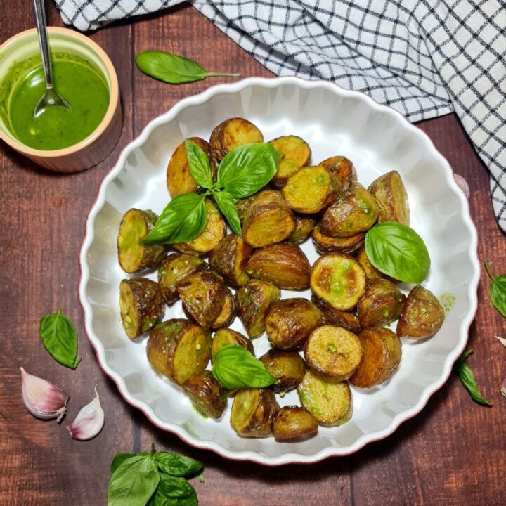 Freshly roasted crispy potatoes garnished with basil on a white scalloped plate, accompanied by a bowl of green pesto sauce.