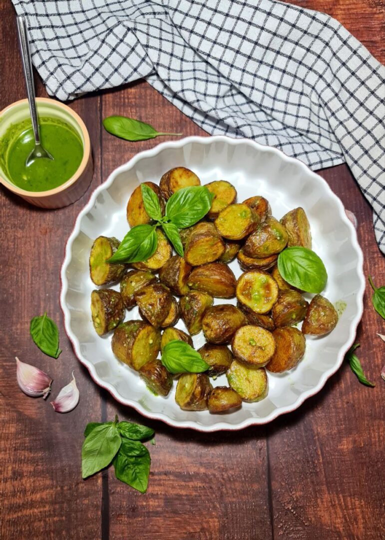 Freshly roasted crispy potatoes garnished with basil on a white scalloped plate, accompanied by a bowl of green pesto sauce.