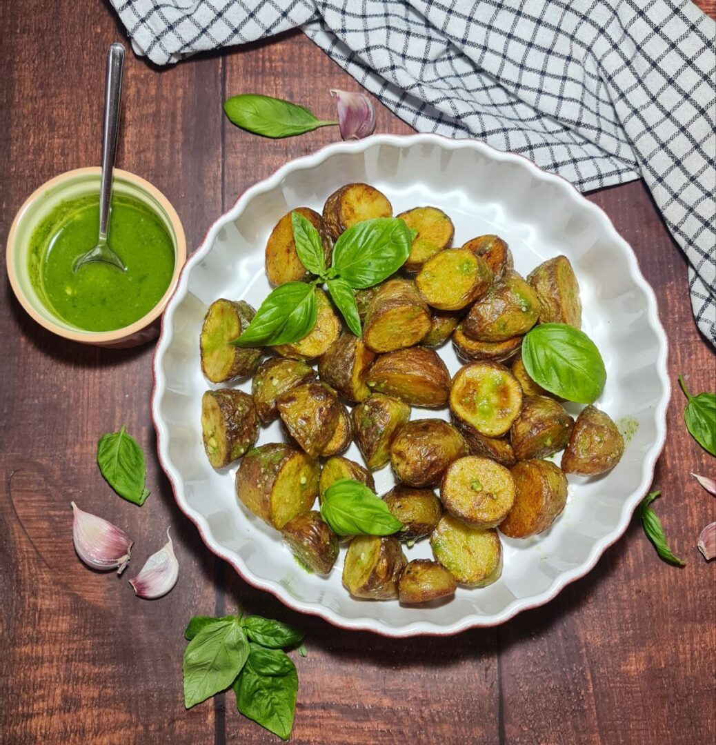 Final presentation of crispy pesto roasted potatoes on a white scalloped plate, garnished with fresh basil, alongside a bowl of green pesto sauce and a rustic kitchen setting.