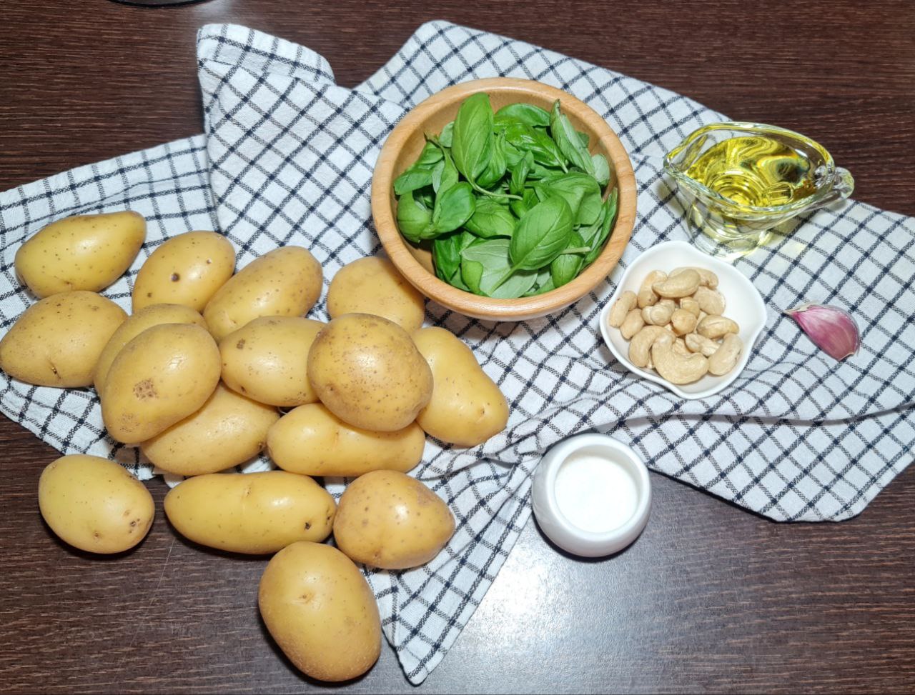 Ingredients for crispy pesto roasted potatoes including fresh basil, potatoes, cashews, garlic, olive oil, and salt on a checkered napkin.