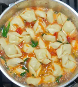 A pot of vegan tortellini soup cooking, with tortellini, spinach, tomatoes, and broth simmering together.