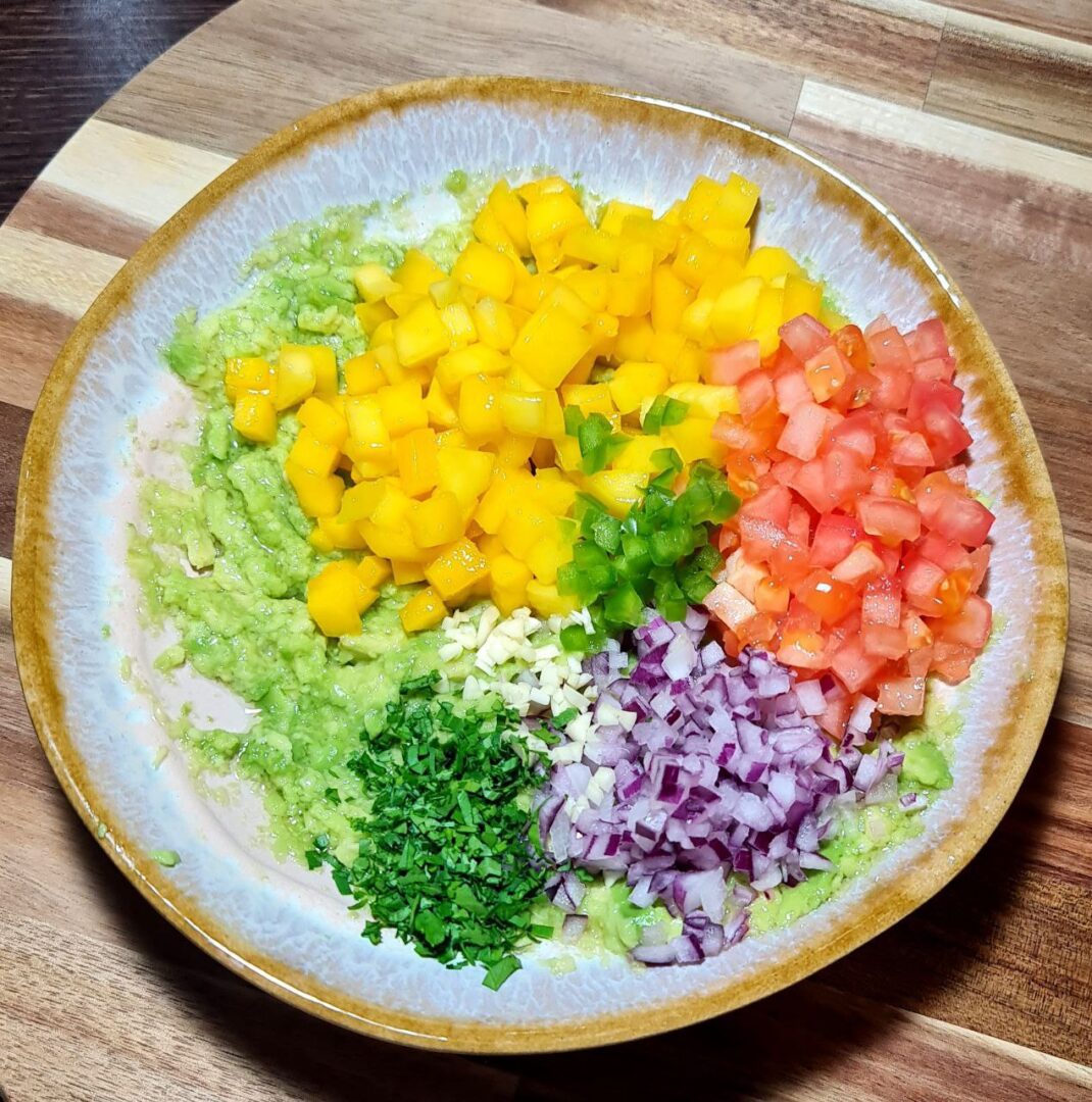  Mango guacamole ingredients in a bowl including mashed avocado, diced mango, red onion, tomato, cilantro, jalapeno, and garlic.