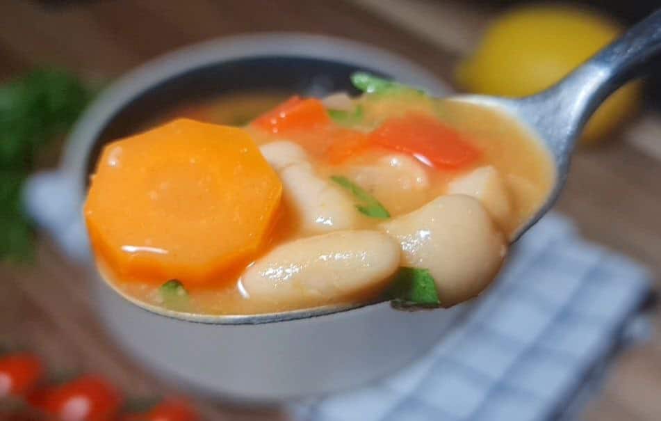 : Close-up of a spoonful of creamy bean soup with carrots, white beans, and red bell peppers.