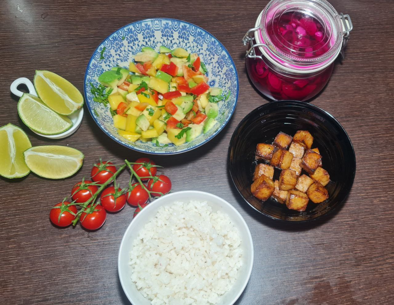 Ingredients for a tropical tofu rice bowl arranged on a wooden table including a bowl of mango salsa, a jar of pickled onions, cherry tomatoes, lime wedges, crispy tofu, and white rice.