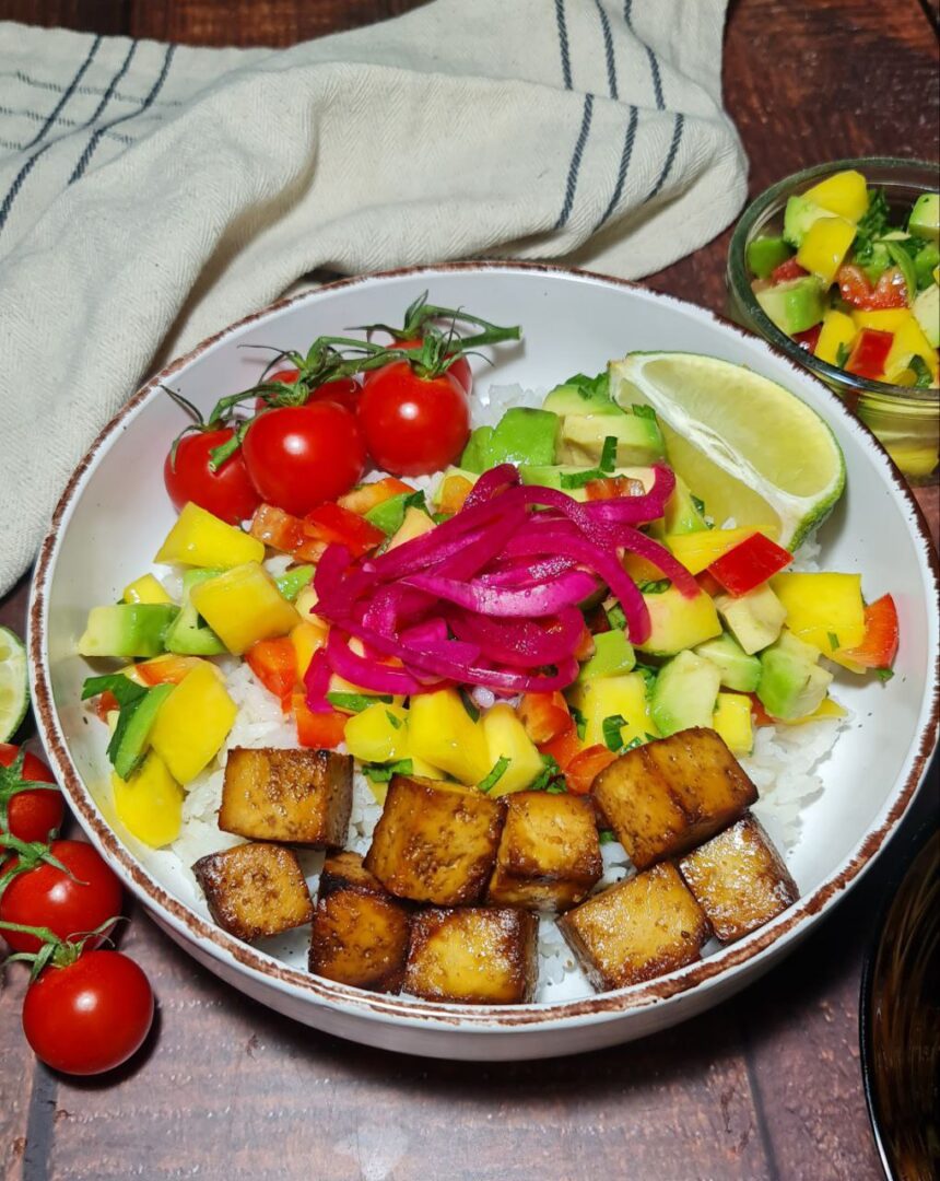 Close-up of a tropical tofu rice bowl with pickled red onions, fresh cherry tomatoes, mango salsa, avocado, and crispy tofu cubes, served with lime wedges on a rustic wooden table.