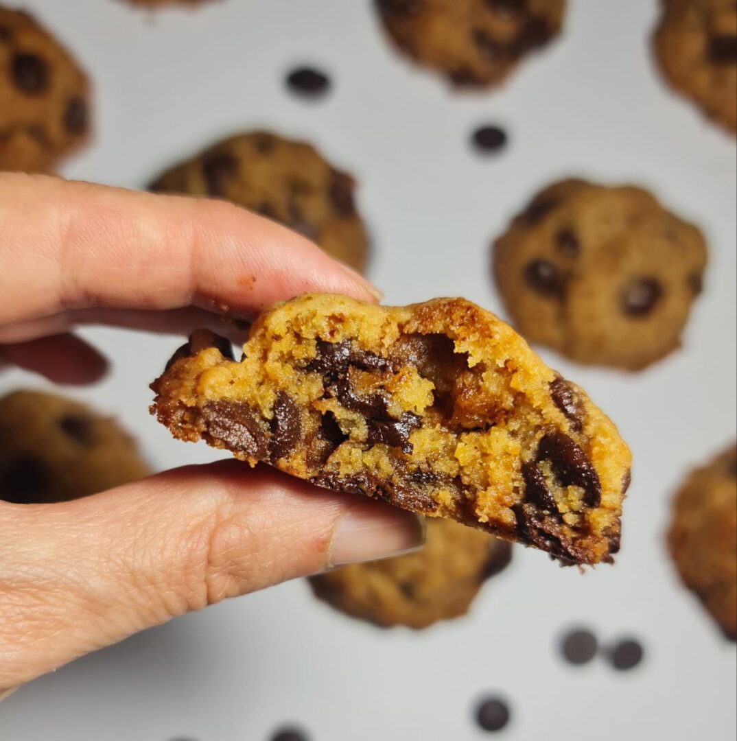 Close-up of a hand holding a half-eaten vegan chocolate chip cookie showing gooey chocolate and a moist, chewy texture, with more cookies in the background