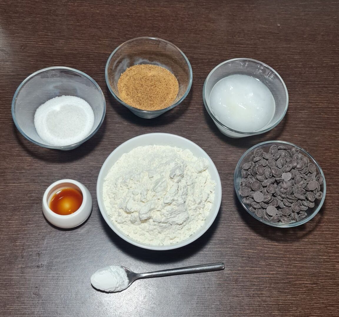 Ingredients for vegan chocolate chip cookies including flour, sugar, brown sugar, coconut oil, vanilla extract, baking soda, and vegan chocolate chips on a wooden table