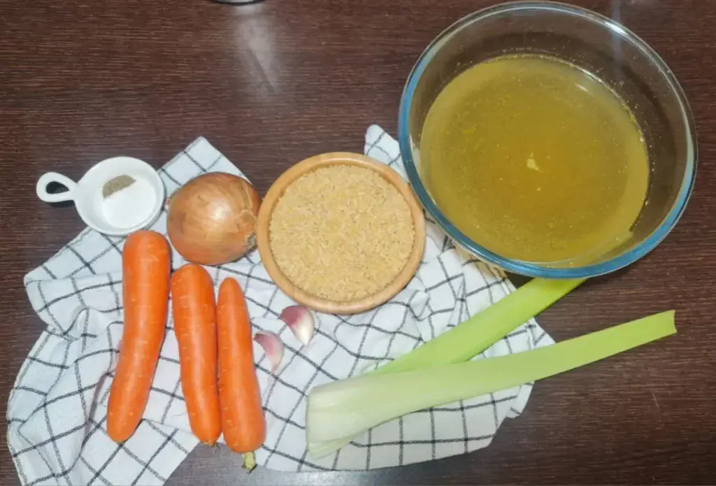 Ingredients for vegan pastina soup including carrots, onion, celery, garlic, orzo pasta, and vegetable broth on a wooden table.