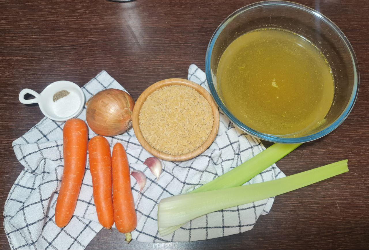  Ingredients for vegan pastina soup including carrots, onion, celery, garlic, orzo pasta, and vegetable broth on a wooden table.