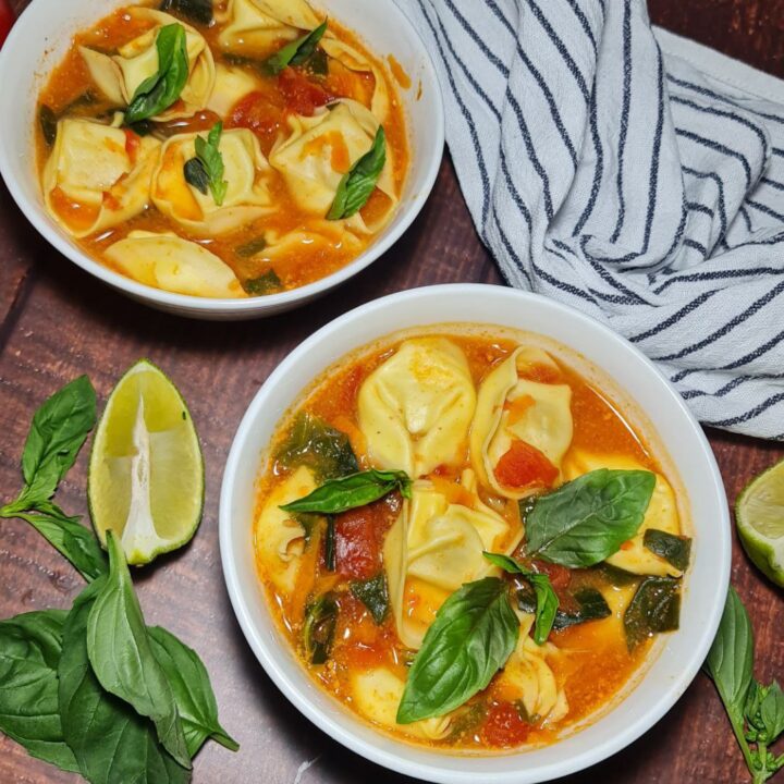 vegan tortellini soup. Two bowls of vegan tortellini soup garnished with fresh basil, served on a wooden table with lime wedges and a striped cloth napkin.