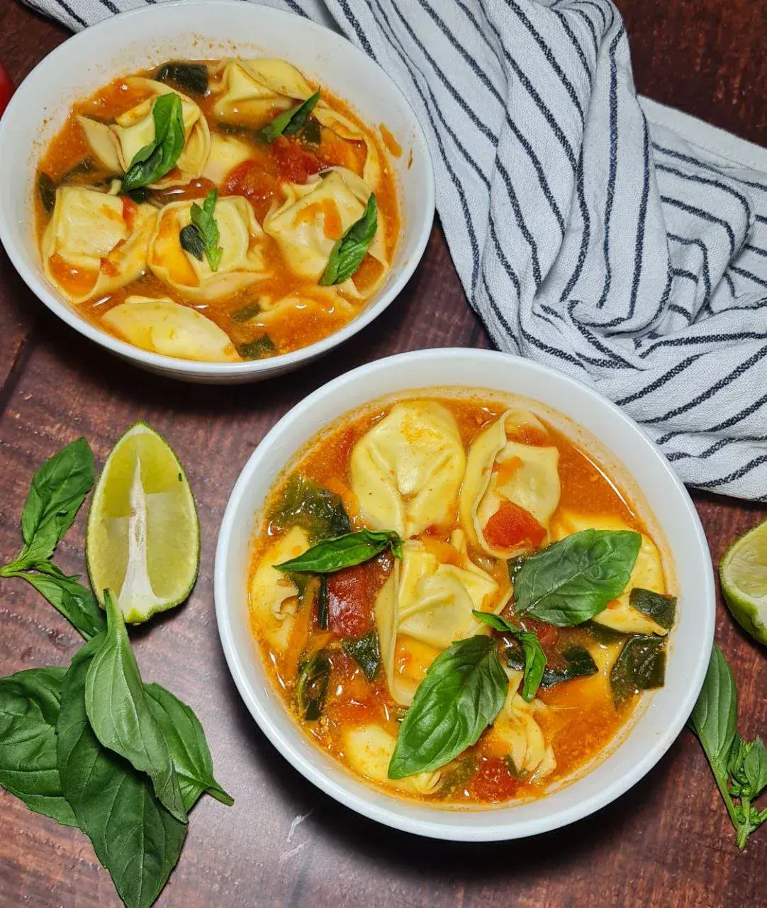 easy vegan tortellini soup. Two bowls of vegan tortellini soup garnished with fresh basil, served on a wooden table with lime wedges and a striped cloth napkin.