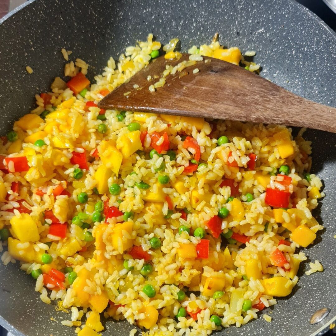  A wok filled with colorful mango fried rice, featuring peas, red bell peppers, and mango chunks, being stirred with a wooden spatula.