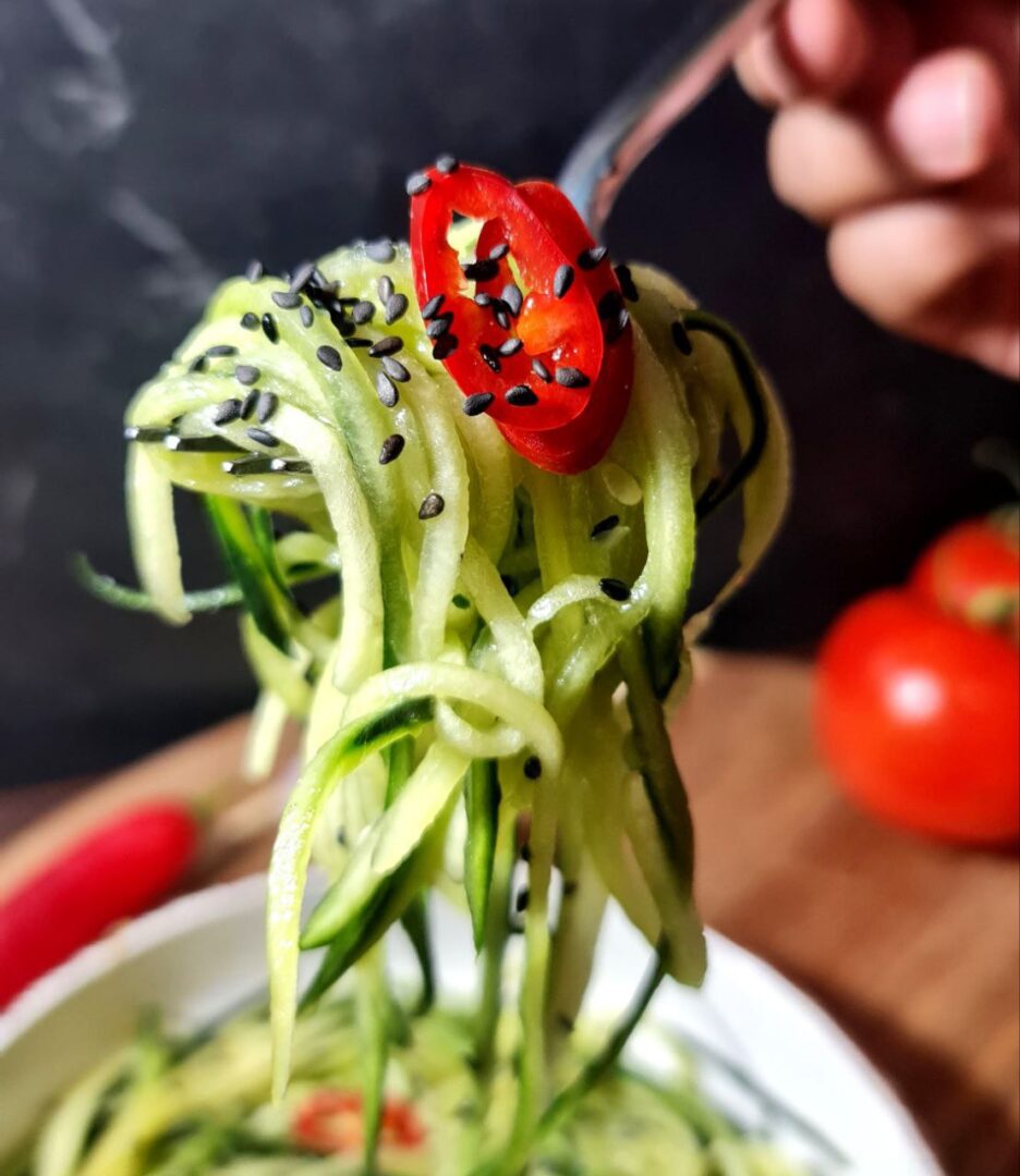  Close-up of a cucumber noodle salad topped with black sesame seeds and a slice of red chili pepper, held by a fork.