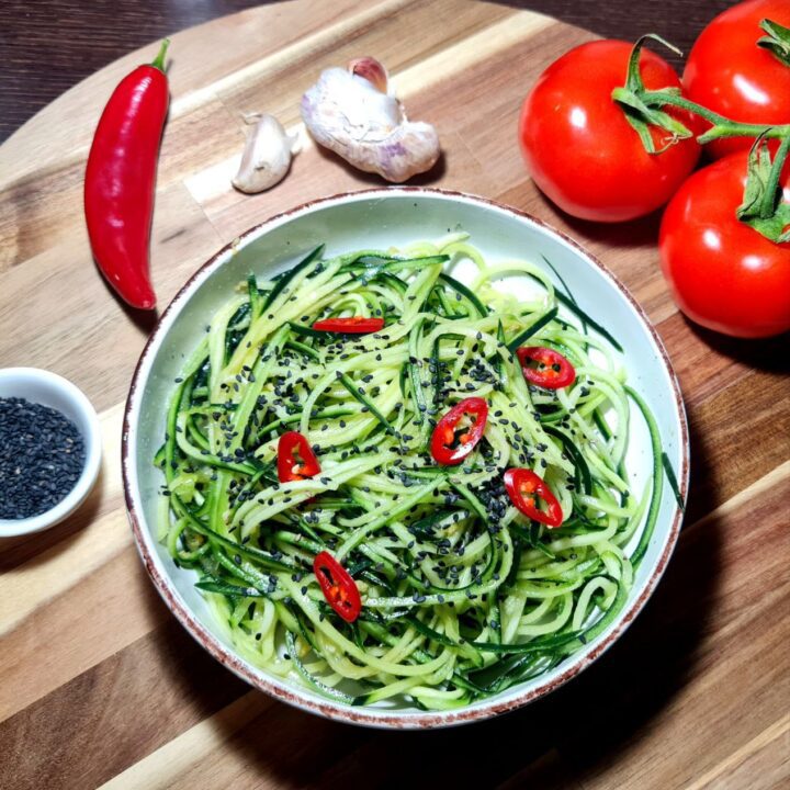 A bowl of cucumber noodle salad garnished with black sesame seeds and red chili slices, surrounded by fresh ingredients including tomatoes, garlic, a chili pepper, and a small bowl of black sesame seeds on a wooden cutting board.