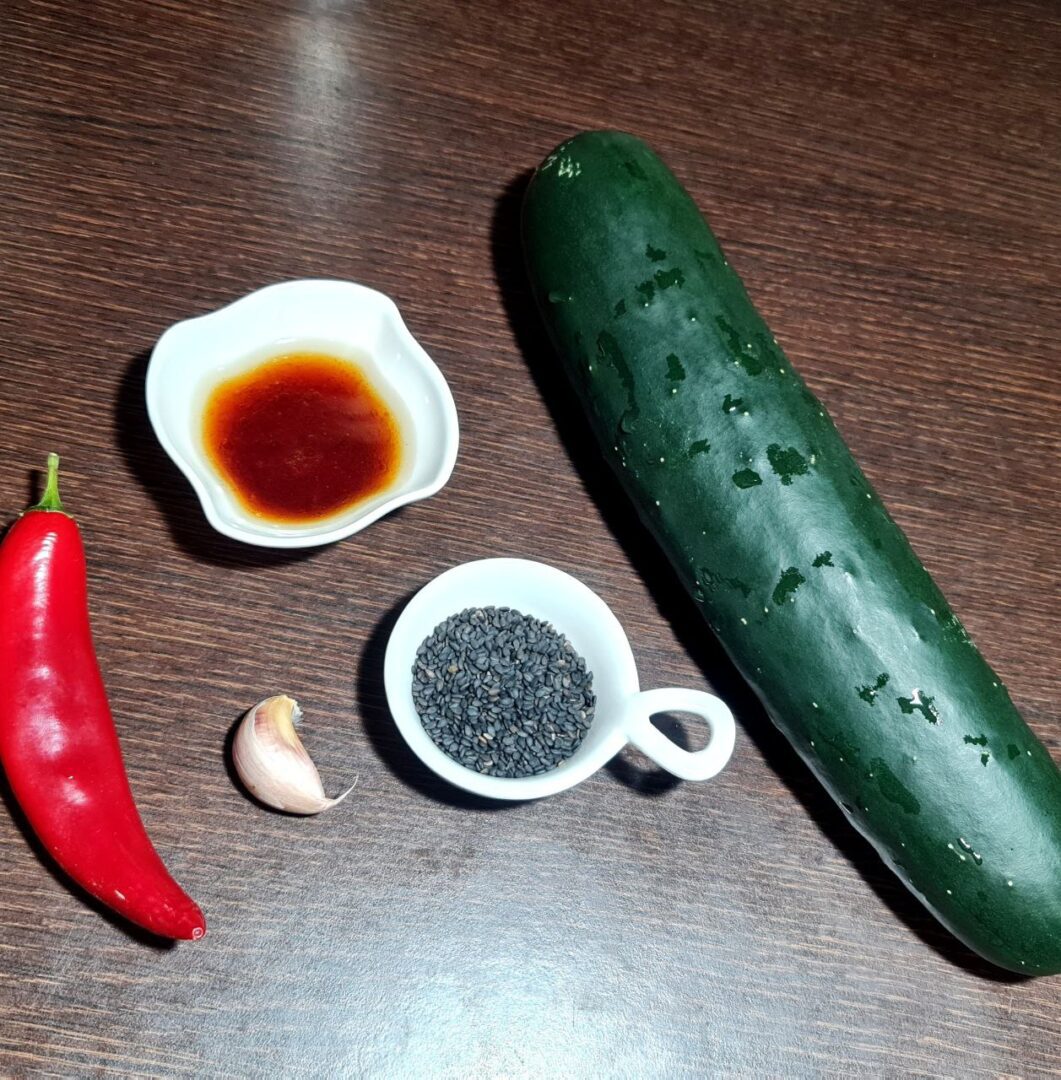  Ingredients for cucumber noodle salad, including a cucumber, red chili pepper, garlic clove, black sesame seeds, and a small dish of sauce, displayed on a wooden surface.