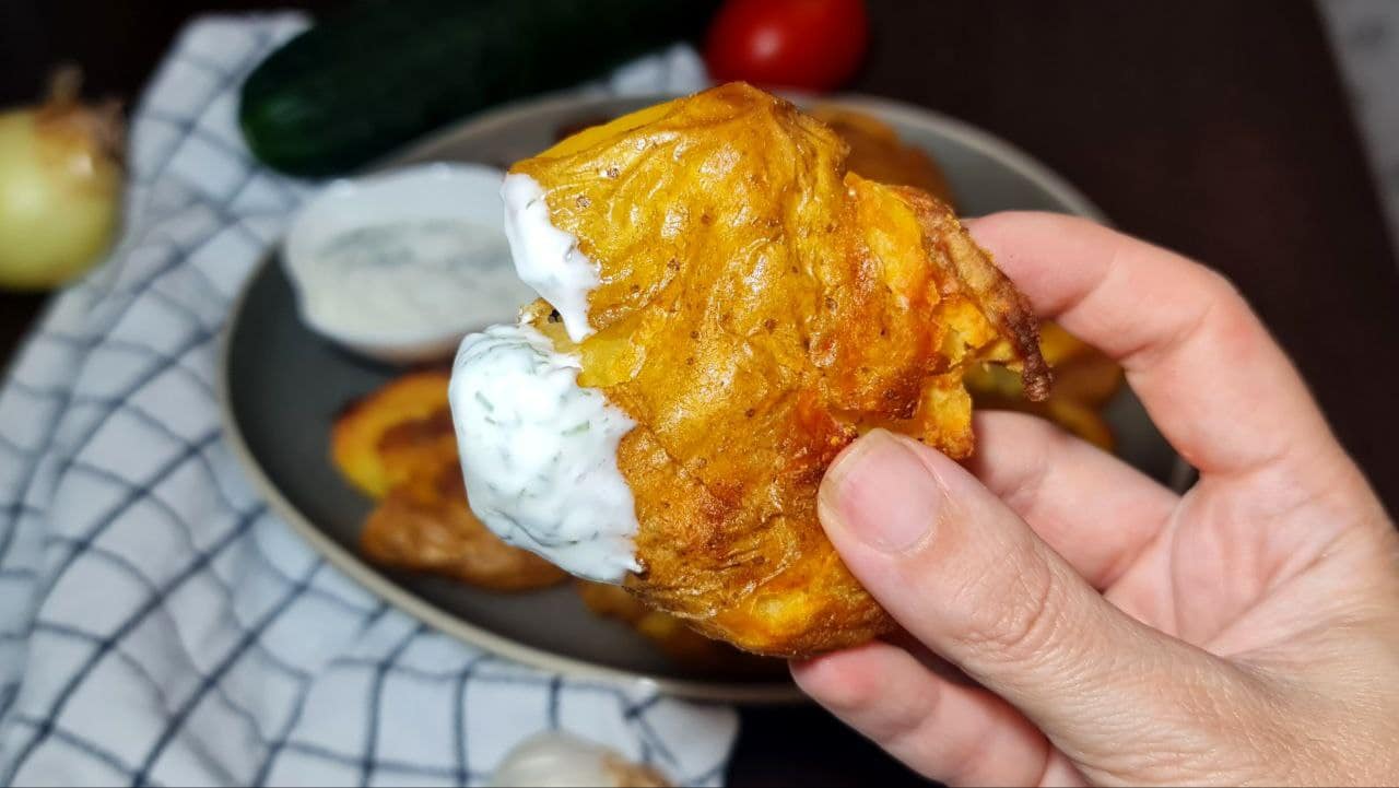 Hand holding a crispy smashed potato dipped in creamy sauce.