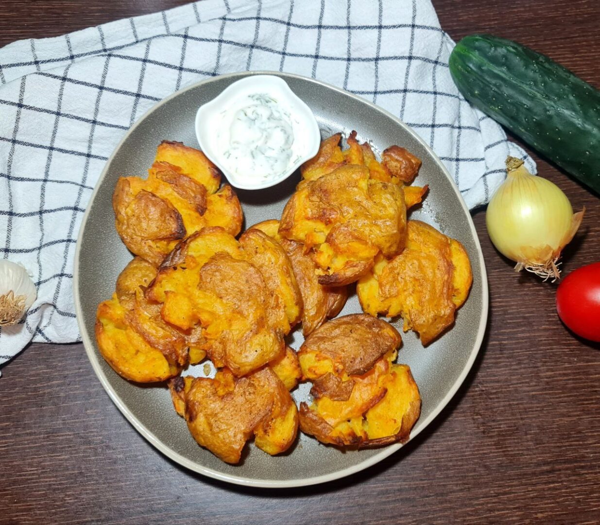  Plate of crispy smashed potatoes with dipping sauce, surrounded by fresh vegetables and cooking ingredients.