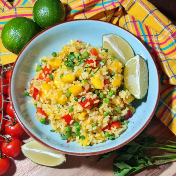 A bowl of vibrant mango fried rice garnished with fresh herbs, accompanied by lime wedges, cherry tomatoes, and a colorful checkered napkin.