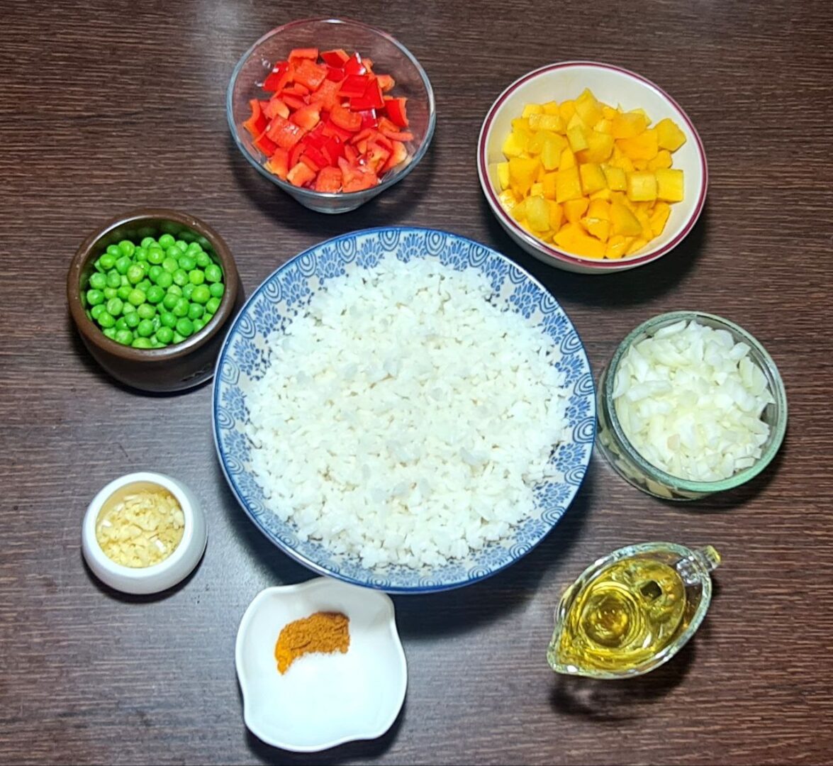 Ingredients for mango fried rice including diced mango, red bell peppers, peas, onions, garlic, spices, oil, and cooked rice, all arranged in bowls on a wooden surface.