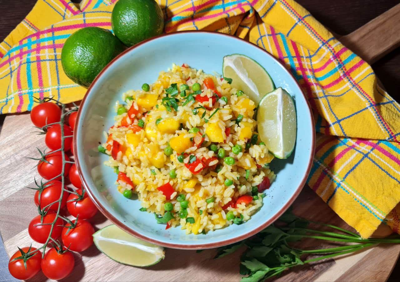 A bowl of vibrant mango fried rice garnished with fresh herbs, accompanied by lime wedges, cherry tomatoes, and a colorful checkered napkin.