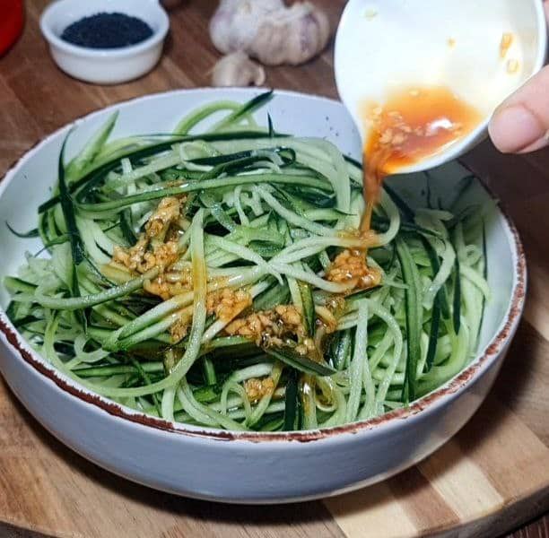 A bowl of cucumber noodle salad being topped with a savory dressing, with black sesame seeds and garlic in the background.