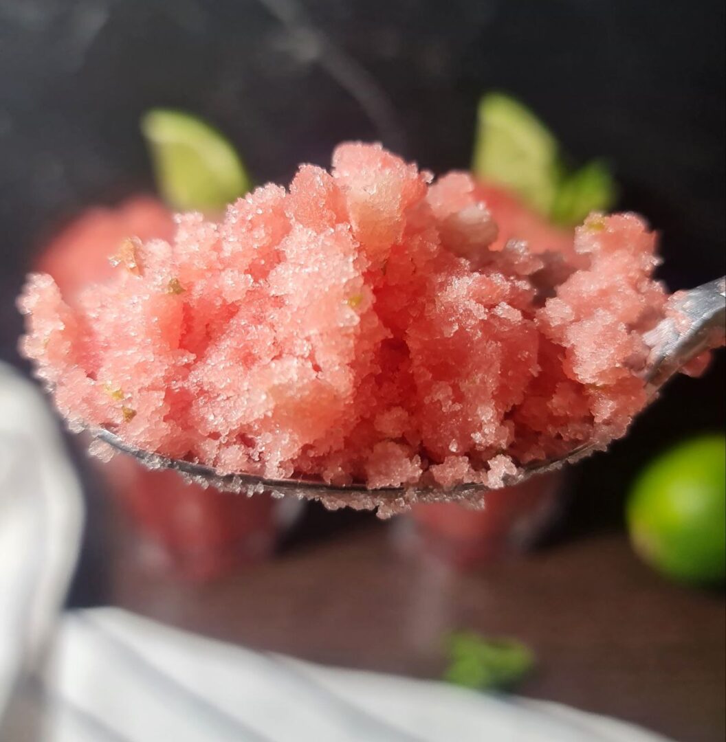 Close-up of a spoonful of refreshing watermelon slushie with a blurred background of watermelon and lime slices.