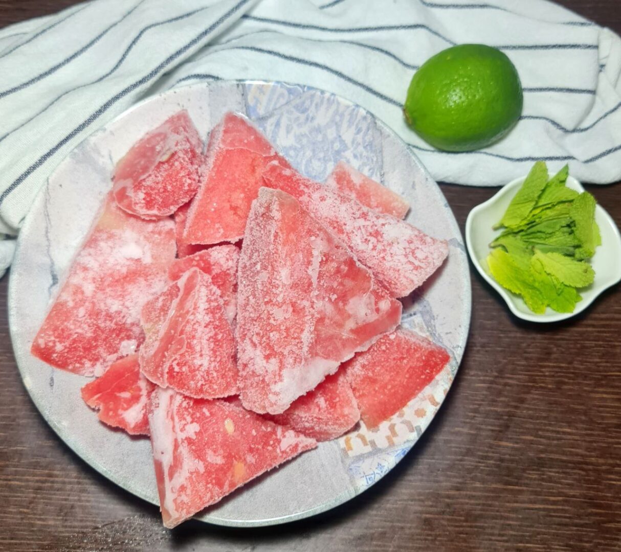  Plate of frozen watermelon pieces with a lime and mint leaves on the side.