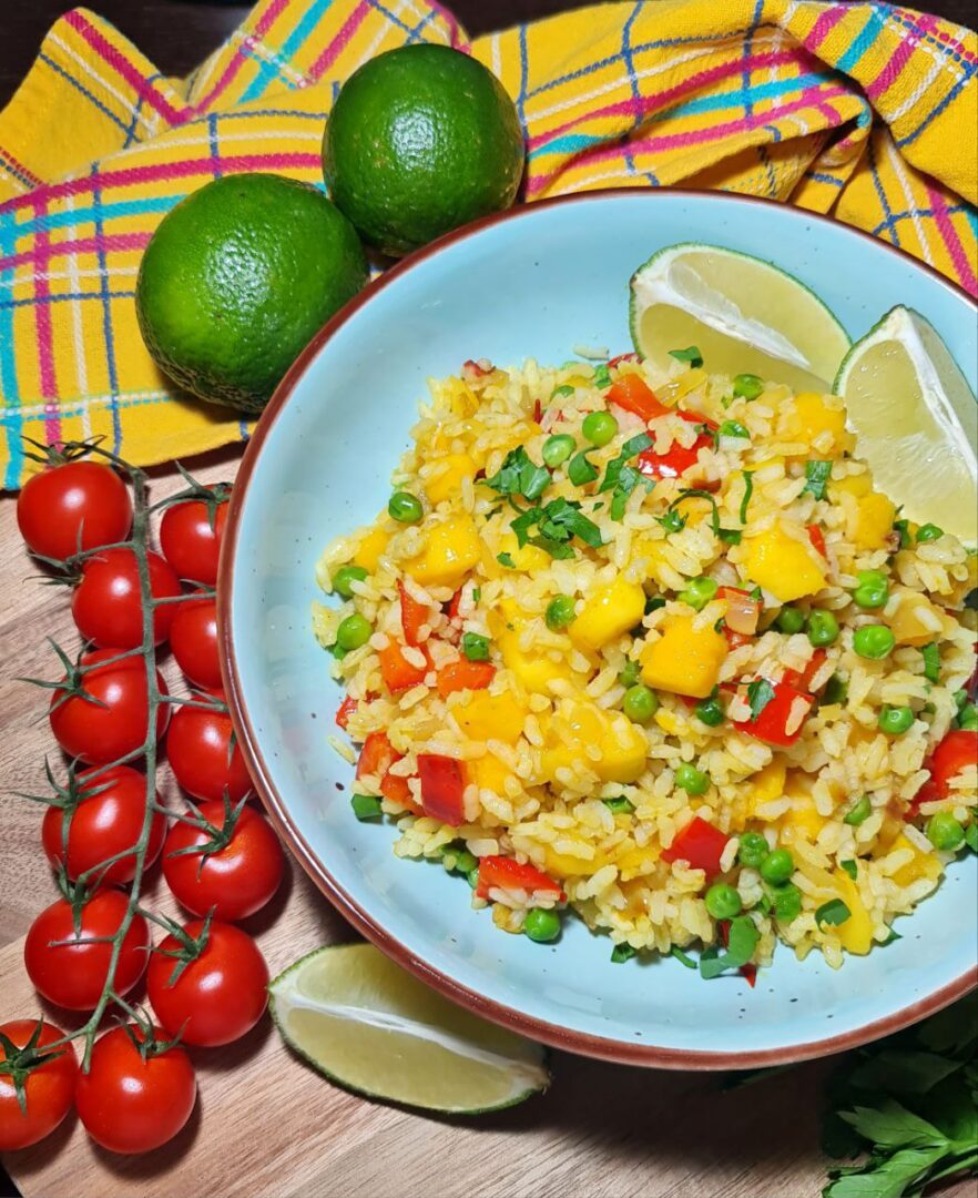  A bowl of vibrant mango fried rice garnished with fresh herbs and lime wedges, surrounded by cherry tomatoes, limes, and a colorful checkered napkin.