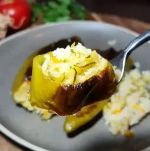 Close-up of a stuffed pepper on a fork with rice and vegetable filling, with a blurred background of a plate and fresh ingredients.