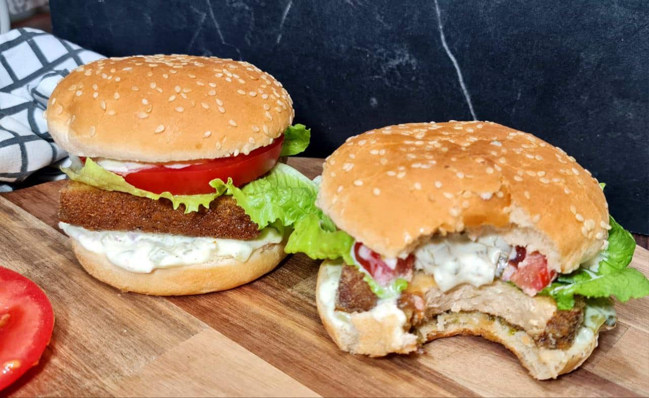 Two vegan tofu burgers, one with a bite taken out, showing the crispy tofu patty, lettuce, tomato, and creamy sauce in a sesame seed bun.