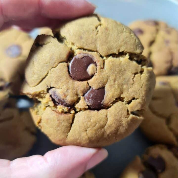 A close-up of a hand holding a vegan matcha chocolate chip cookie, showcasing its cracked surface and chocolate chips.