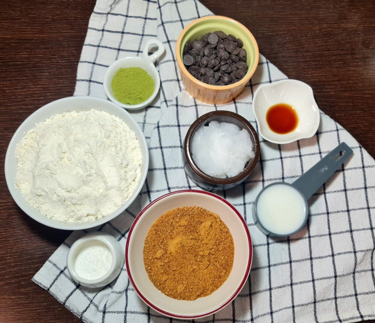  Ingredients for vegan matcha chocolate chip cookies displayed on a checkered cloth, including flour, matcha powder, chocolate chips, coconut oil, brown sugar, vanilla extract, and plant-based milk.