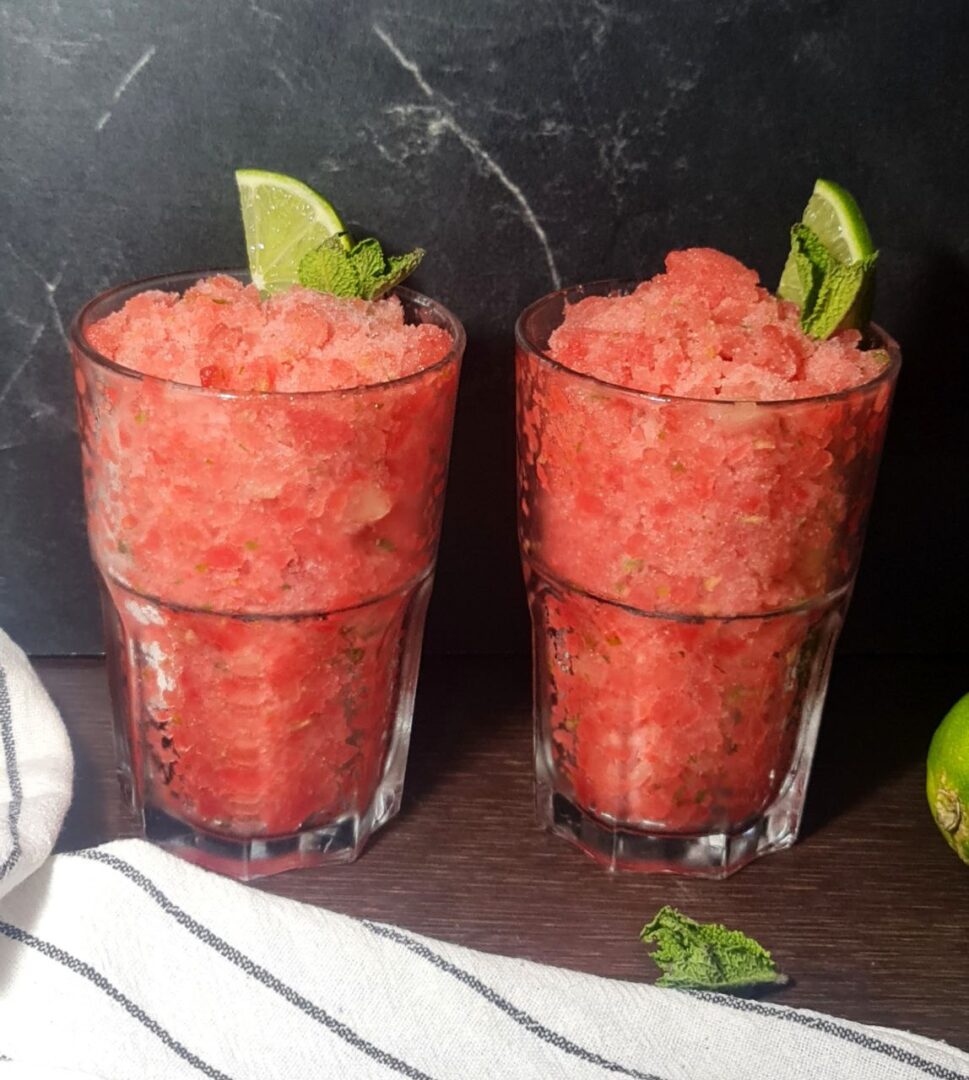 Two glasses of refreshing watermelon slushie garnished with lime wedges and mint leaves, with whole and cut limes beside them.