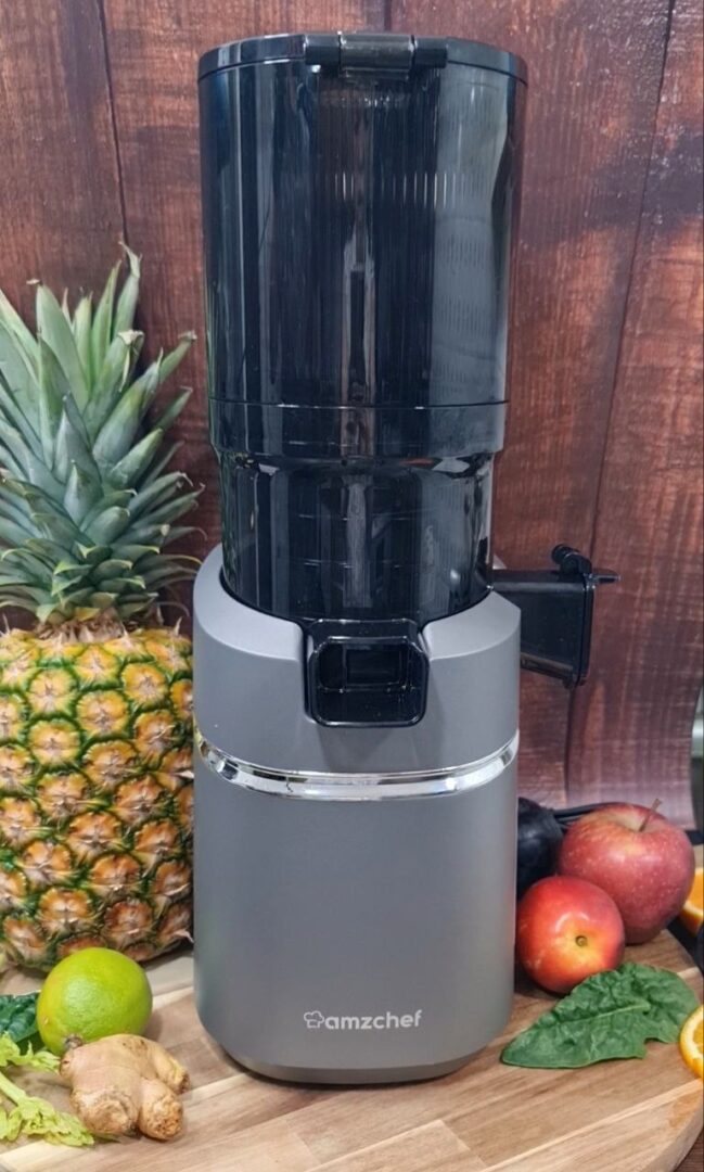 A sleek AMZCHEF juicer surrounded by fresh fruits and vegetables including pineapple, lime, ginger, apples, and leafy greens.