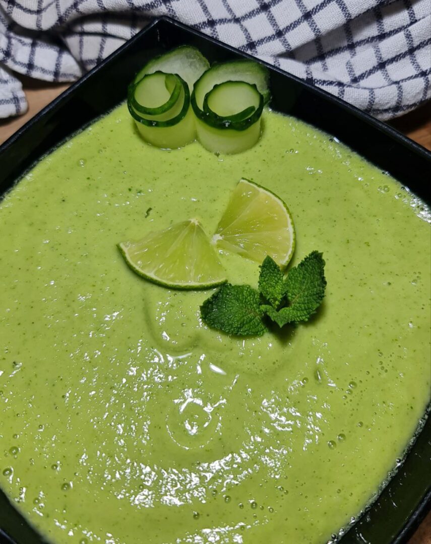 Close-up view of cold avocado cucumber soup garnished with lime wedges, cucumber rolls, and mint leaves.