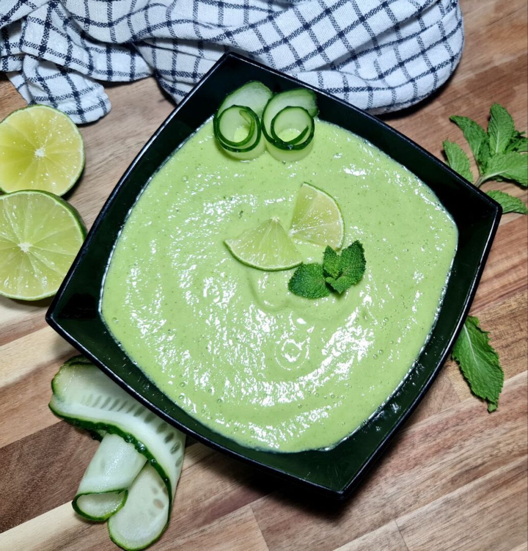 A refreshing bowl of cold avocado cucumber soup garnished with lime wedges, cucumber slices, and mint leaves.