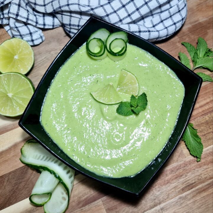 tA refreshing bowl of cold avocado cucumber soup garnished with lime wedges, cucumber slices, and mint leaves.