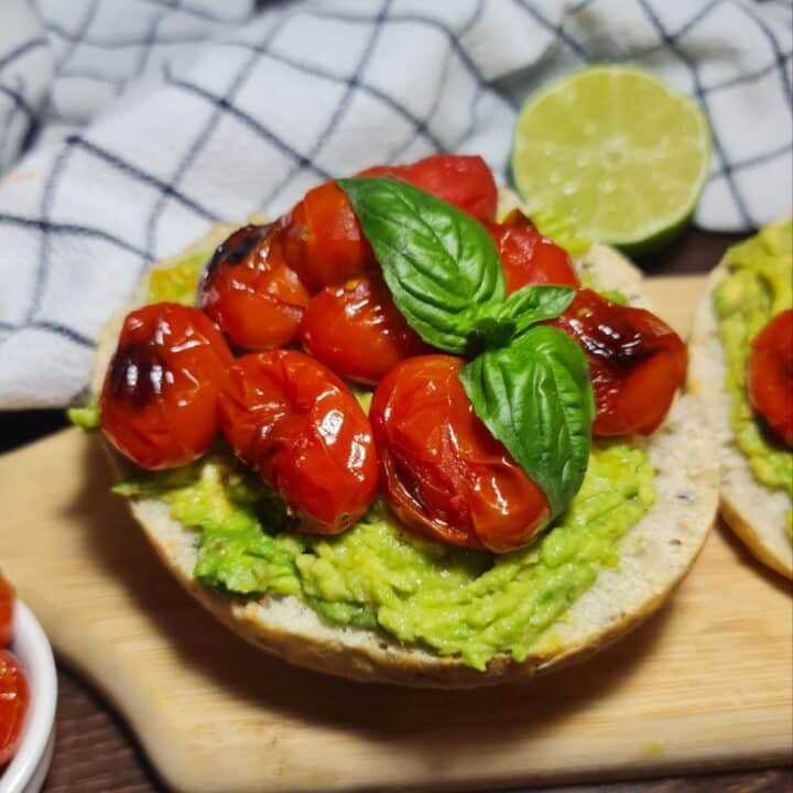 Delicious vegan avocado toast topped with roasted cherry tomatoes and fresh basil, served on a wooden board with a sliced lime in the background. Easy vegan avocado toast topped with roasted cherry tomatoes and fresh basil, perfect for a healthy breakfast.