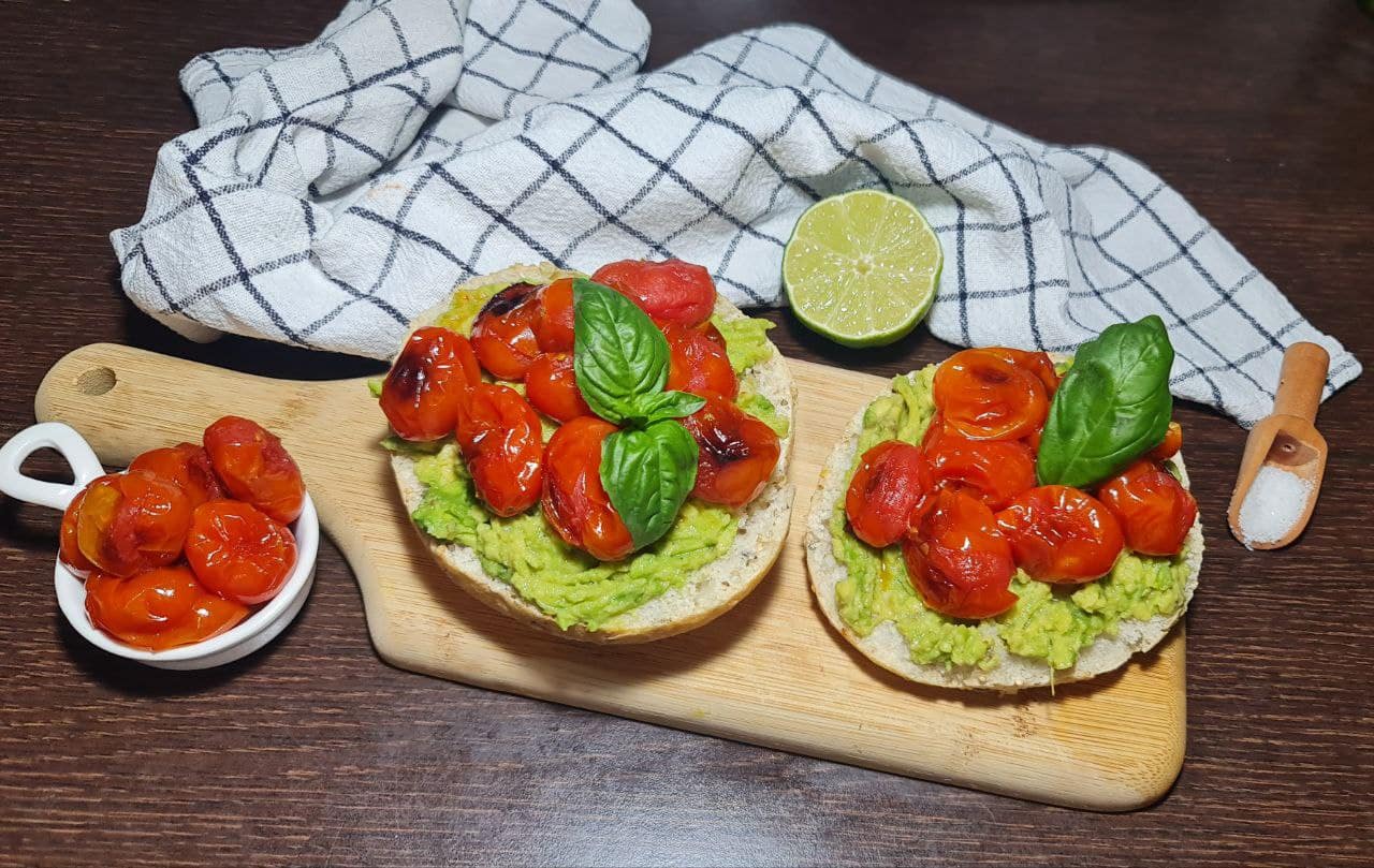 Vegan avocado toast topped with roasted cherry tomatoes and fresh basil, served on a wooden board with a half lime and a bowl of cherry tomatoes on the side. Easy vegan avocado toast topped with roasted cherry tomatoes and fresh basil, served with a side of extra tomatoes and lime.