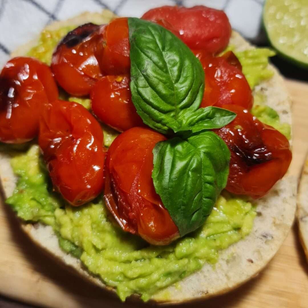  Easy Vegan avocado toast recipe for breakfast with roasted cherry tomatoes and fresh basil leaves, presented on a wooden board with a halved lime and extra tomatoes on the side. Vegan avocado toast with roasted cherry tomatoes and fresh basil, accompanied by a lime and extra tomatoes.