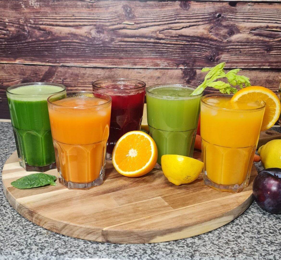 A colorful assortment of five freshly made juices, featuring green, orange, and red varieties, garnished with fruits and vegetables.