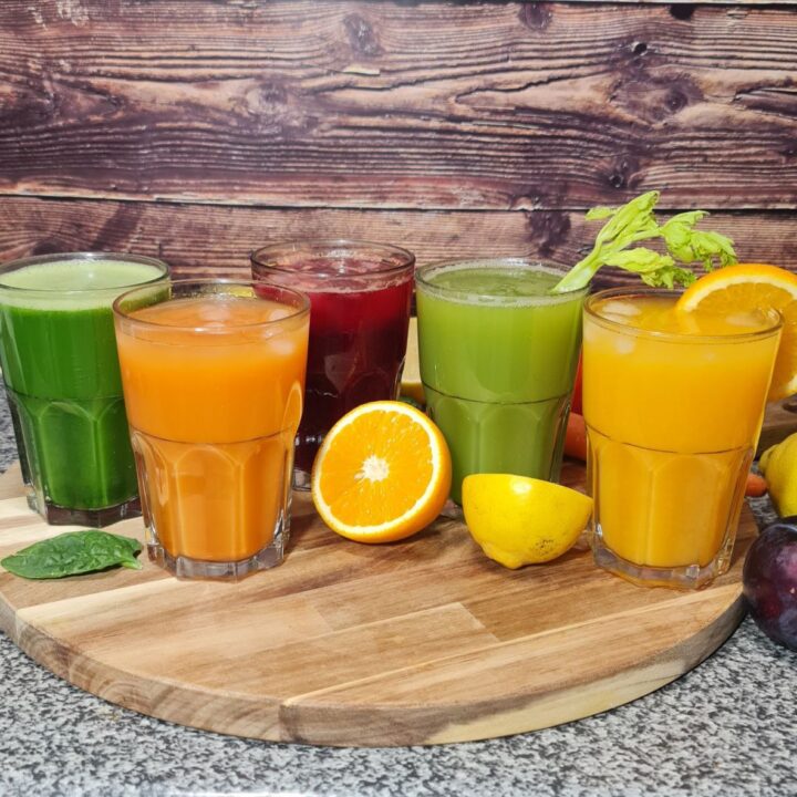 A colorful assortment of five freshly made juices, featuring green, orange, and red varieties, garnished with fruits and vegetables.